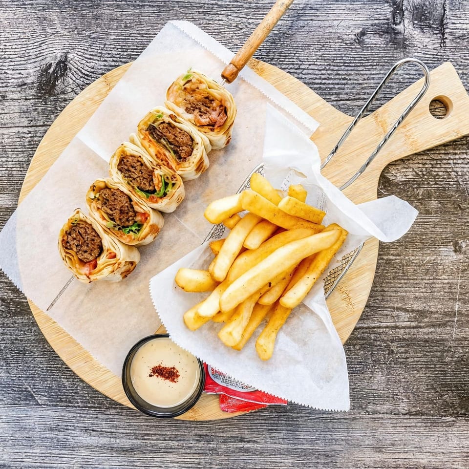 Sliced beef wraps with lettuce and tomato served alongside golden fries and dipping sauce on a wooden board.
